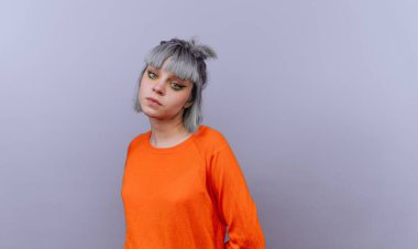 Gri saçlı, turuncu süveterli, gri arka planda genç bir kadın..