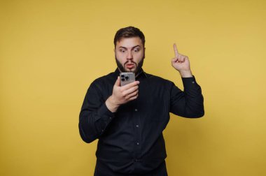 Siyah gömlekli adam akıllı telefona şaşırmış, sarı arka planda..