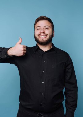 Siyah gömlekli, gülümseyen adam mavi arka planda başparmak işareti yapıyor.