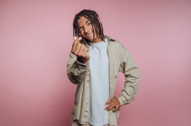Young man with braided hair making a heart gesture against pink background.