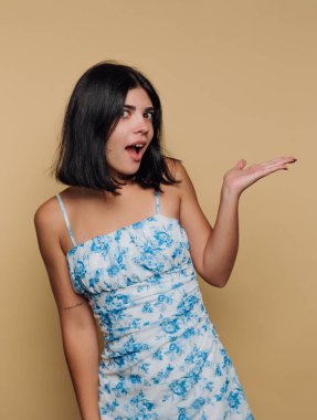 Omuz hizasında siyah saçlı, çiçekli bir elbise giyen, bej arka planda şakacı poz veren genç bir kadın.