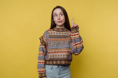 Renkli desenli süveterli bir kadın, sarı arka planda düşünceli bir şekilde el kol hareketi yapar..