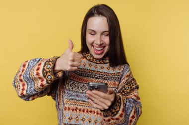 Süveter giymiş bir kadın telefona bakarken başparmağını kaldırıyor.