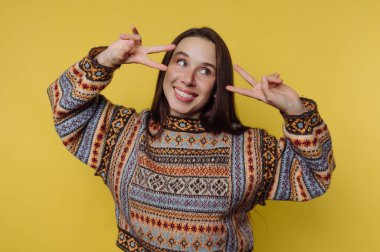 Sarı arka planda barış işaretleri yapan desenli süveterli gülümseyen kadın..