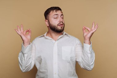 Beyaz gömlekli adam bej arka planda gözleri kapalı meditasyon yapıyor..