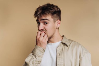 Tırnak yiyen genç bir adam, bej arka planda endişeli görünüyor..