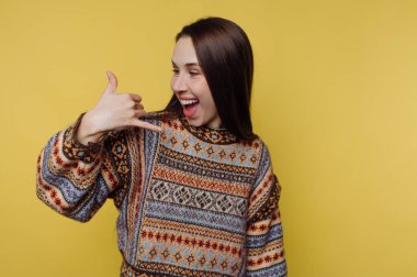 Süveter giymiş gülümseyen bir kadın sarı arka planda bir telefon hareketi yapıyor..