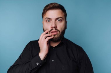 Siyah gömlekli adam mavi arka planda şaşırmış bir ifade var.