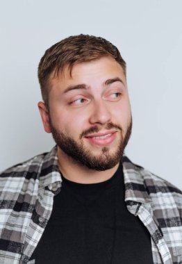 A young man with a beard wearing a plaid shirt and black t-shirt, looking to the side with a playful expression