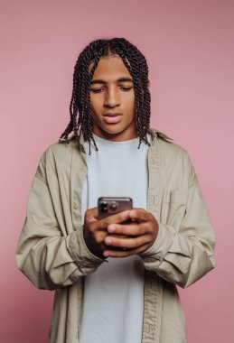 Saçları örgülü genç bir adam pembe arka planda akıllı telefon kullanıyor..