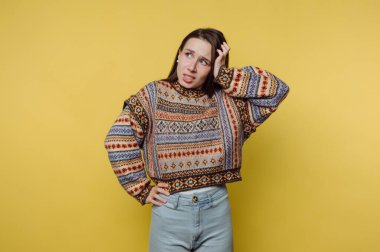 Renkli kazaklı bir kadın sarı arka planda duruyor, kafası karışık görünüyor..