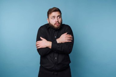 Siyah gömlekli adam üşümüş, kollarını kavuşturmuş, mavi arka plan.