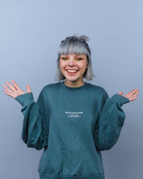 Young woman with gray hair and bangs, wearing a teal sweatshirt, smiling with arms raised against a plain blue background.