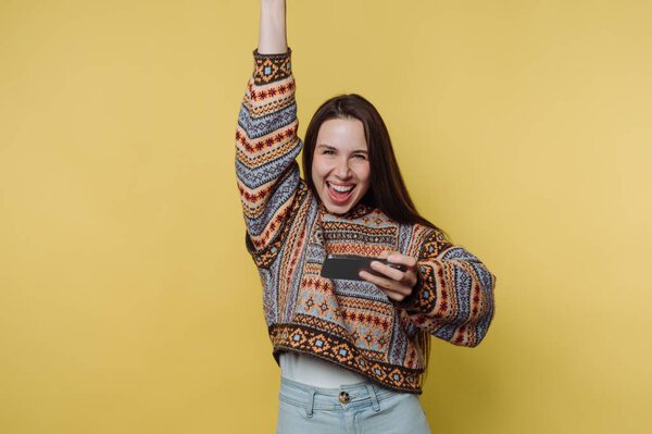 A joyful young woman in a colorful sweater holding a smartphone, celebrating with one arm raised against a yellow background