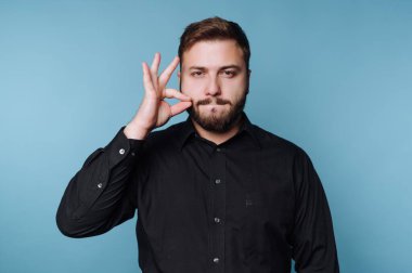 Bir adam mavi bir duvara karşı duruyor ve eli çenesinin yanında poz veriyor. Bir jest yaparken odaklanmış görünüyor. Ayarları basit ve hafif..