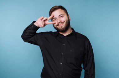Bir adam mavi arka planda duruyor ve eliyle barış işareti yaparken gülümsüyor. Siyah bir gömlek giyiyor ve anın tadını çıkarıyor gibi görünüyor..