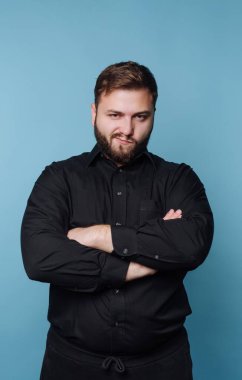 Sakallı bir adam kollarını kavuşturmuş ve yüzünde ciddi bir ifadeyle ayakta duruyor. Siyah bir gömlek giyer ve düz mavi bir duvara karşı poz verir..