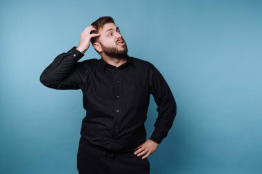 Düşünceli bir ifadeyle duran ve başını kaşıyan bir adam. Siyah bir gömlek giyiyor ve biraz kafası karışmış görünüyor. Arkaplan koyu mavi renkte..