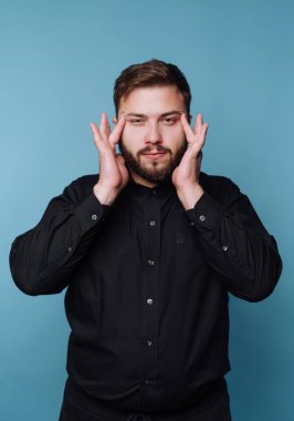 Genç bir adam mavi bir duvarın karşısında dururken iki eliyle yüzünü tutuyor. O derin düşüncelere dalmış gibi görünüyor. Ayarlar rahat ve resmi görünmüyor..