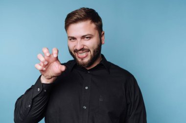 Bir adam mavi bir arka planın önünde duruyor, gülümserken neşeli bir el hareketi gösteriyor. Siyah bir gömlek giyiyor ve eğlenceli bir anın tadını çıkarıyor gibi görünüyor..