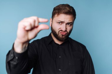 Bir adam mavi bir arkaplanın önünde duruyor ve küçük bir miktar belirtmek için elleriyle bir jest yapıyor. Yüzündeki ifade, poz verirken neşeli bir ruh hali gösteriyor..