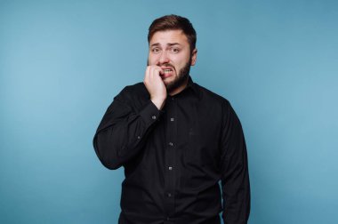 Bir adam tırnaklarını yerken mavi bir duvara dayanıyor. Yüzünde gerginlik belirtileri var ve endişeli görünüyor. Bu sahne gündüz vakti kapalı bir yerde geçiyor..