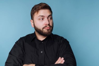 Bir adam mavi bir duvarın önünde duruyor, kolları bağlı ve yana bakıyor. Sakalı var ve siyah bir gömlek giyiyor. İçerisi yumuşak ışıklandırmayla dolu..