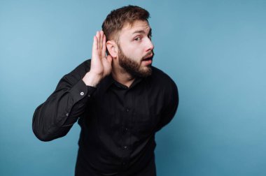 Mavi arka planda duran siyah gömlekli bir adam. Elini kulağına dayayıp bir şey duymaya çalıştığını gösteriyor. İfadesi ilgi gösteriyor..