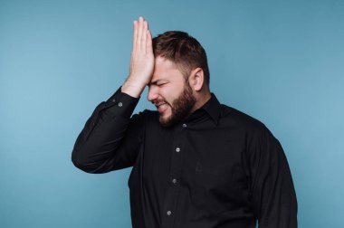 Bir adam mavi bir arka plana karşı duruyor ve hayal kırıklığına uğramış bir ifadeyi gösterirken elini alnına koyuyor. Siyah bir gömlek giyiyor ve derin düşüncelere dalmış gibi görünüyor..