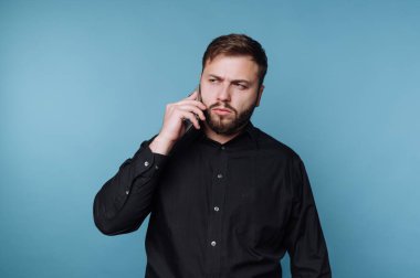 Bir adam kulağına telefon tutarken mavi bir duvara dayanıyor. Odaklanmış görünüyor ve siyah bir gömlek giyiyor. Işıklandırma yüz ifadesini vurguluyor..