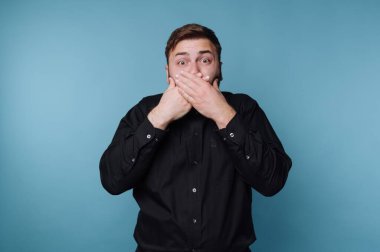Kısa kahverengi saçlı bir adam mavi arka planda duruyor. İki eliyle ağzını kapatıyor ve şaşkınlık ifadesi gösteriyor. Onun siyah gömleği arka planla çelişiyor..