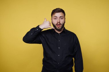 Bir adam sarı bir duvarın önünde şaşırmış bir ifadeyle duruyor. Bir telefon konuşmasını taklit etmek için elini kulağına dayar. Ayar parlak ve neşeli görünüyor..