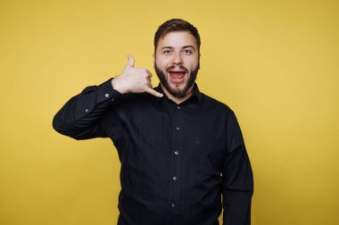 Bir adam sarı bir duvarın önünde duruyor ve eliyle bir jest yaparken gülümsüyor. Siyah bir gömlek giyiyor ve bu eğlenceli ortamda mutlu görünüyor..