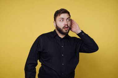 Siyah gömlekli bir adam eli kulağında, şaşırmış bir şekilde duruyor. Sarı arka planlı bir stüdyoda ve dinlemeye odaklanmış gibi görünüyor..