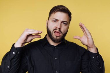 Siyah gömlekli bir adam sarı bir duvarın önünde duruyor. Konuşurken ellerini kullanarak ne demek istediğini açıklıyor veya vurguluyor. Aydınlık ve canlı bir ortam..