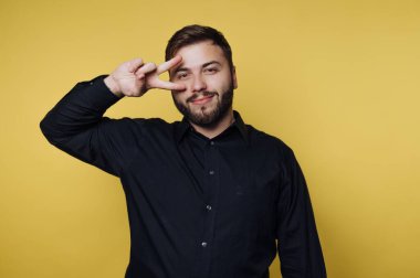 Bir adam sarı bir arka plana karşı duruyor, neşeli bir gülümseme ve bir barış işareti tutuyor. Siyah bir gömlek giyiyor ve kameraya poz verirken cana yakın görünüyor..