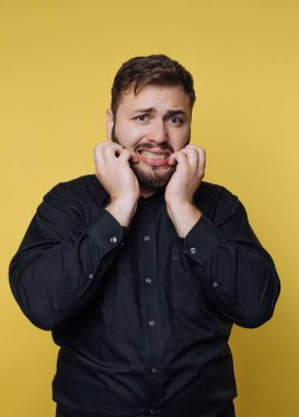 Bir adam yüzünde endişeli bir ifadeyle ayakta duruyor, tırnaklarını yiyor ve ellerini tutuyor. Siyah bir gömlek giymiş ve parlak sarı bir duvara karşı poz veriyor..