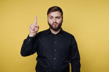 Bir adam sarı bir arkaplanın önünde duruyor. İşaret parmağını kaldırıyor ve yüz ifadesi takınıyor. Siyah bir gömlek giyiyor ve evde dikilirken ciddi görünüyor..