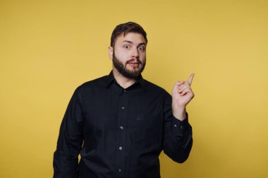 Siyah gömlekli bir adam sarı arka planda duruyor. Tek eliyle işaret ediyor ve şaşırmış görünüyor. Onun ifadesi heyecan ya da merak gösteriyor..