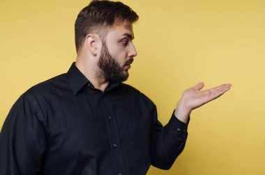 Siyah gömlekli bir adam, yüzünü yana doğru çevirirken eli açık hareket eder. Parlak sarı arka plan sahneye bir renk katıyor. Bir sohbete katılmış gibi görünüyor..