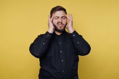 Elleri başının üstünde, yüzünü buruşturan bir adam. Hayal kırıklığı ya da rahatsızlık hissediyor gibi görünüyor. Arka plan sarı ve parlak. Stüdyo ortamını gösteriyor..