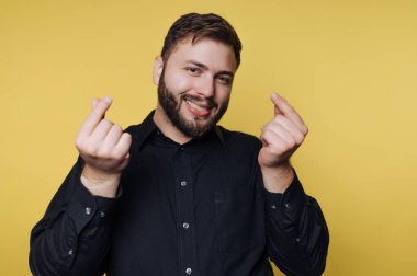 Sarı bir arkaplanın önünde neşeli bir ifadeyle duran bir adam. Genellikle sevgiyle ilişkilendirilen bir el hareketi gösteriyor. Gündüz vakti ve o koyu renk bir gömlek giyiyor..