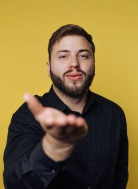 Genç bir adam basit bir iç mekanda duruyor. Doğrudan kameraya bakarken elini kameraya doğru uzatıyor. Arka plan parlak sarı, sahneye neşeli bir dokunuş katıyor..