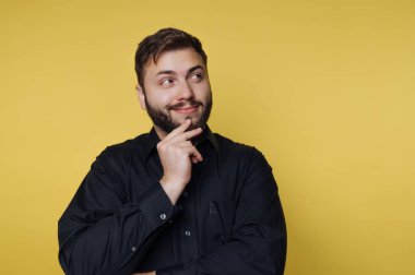 Bir adam hafif bir gülümseme ile ayakta duruyor, yana bakıyor. Sakalı var ve siyah bir gömlek giyiyor. Arka plan sarı, parlak bir ortam yaratıyor..