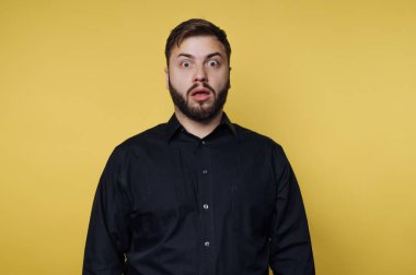 Bir insan şaşırmış bir ifadeyle, geniş gözlerle ve açık ağızla ayakta durur. Arka plan parlak sarı ve kişi siyah bir gömlek giyiyor. Bu sahne güçlü bir tepki veriyor..