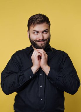 Sarı bir arkaplanın önünde neşeli bir ifadeyle gülümseyen bir adam. Siyah bir gömlek giyiyor ve eğlenceli ya da sinsi bir şeyler düşünüyor gibi görünüyor..