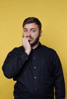 Bir adam gergin bir ifadeyle parlak sarı bir arka plana karşı duruyor. Tırnaklarını yiyor ve şaşırmış görünüyor. Günlük kıyafetleri sahnenin rahatlatıcı havasını arttırıyor..