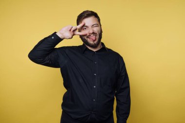 Sarı bir duvarın önünde genç bir adam duruyor. Komik bir ifade takınıyor ve parmaklarıyla barış işareti gösteriyor. İçerideki anın tadını çıkarıyor gibi..