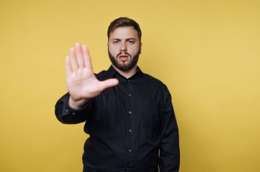 Sarı bir duvarın önünde duran bir adam dur işareti olarak elini kaldırır ve net bir mesaj verir. Siyah bir gömlek giyiyor ve doğrudan kameraya bakıyor..