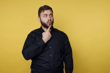 Sarı arka planda bir adam duruyor. Sakalı var ve siyah bir gömlek giyiyor. Yanlara bakarken çenesine dokunuyor, düşüncelere dalmış..